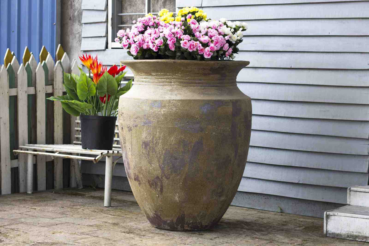 Large antique-finish ceramic urn planter with pink and white flowers, beside a house with a white picket fence.