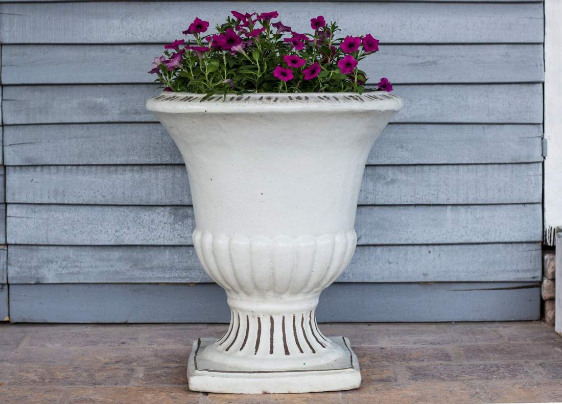 White fluted pedestal urn planter with square base, planted with magenta flowers, against gray wood siding.