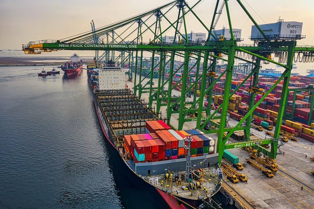 An aerial view of a busy container port with a large ship being unloaded by several towering green cranes.