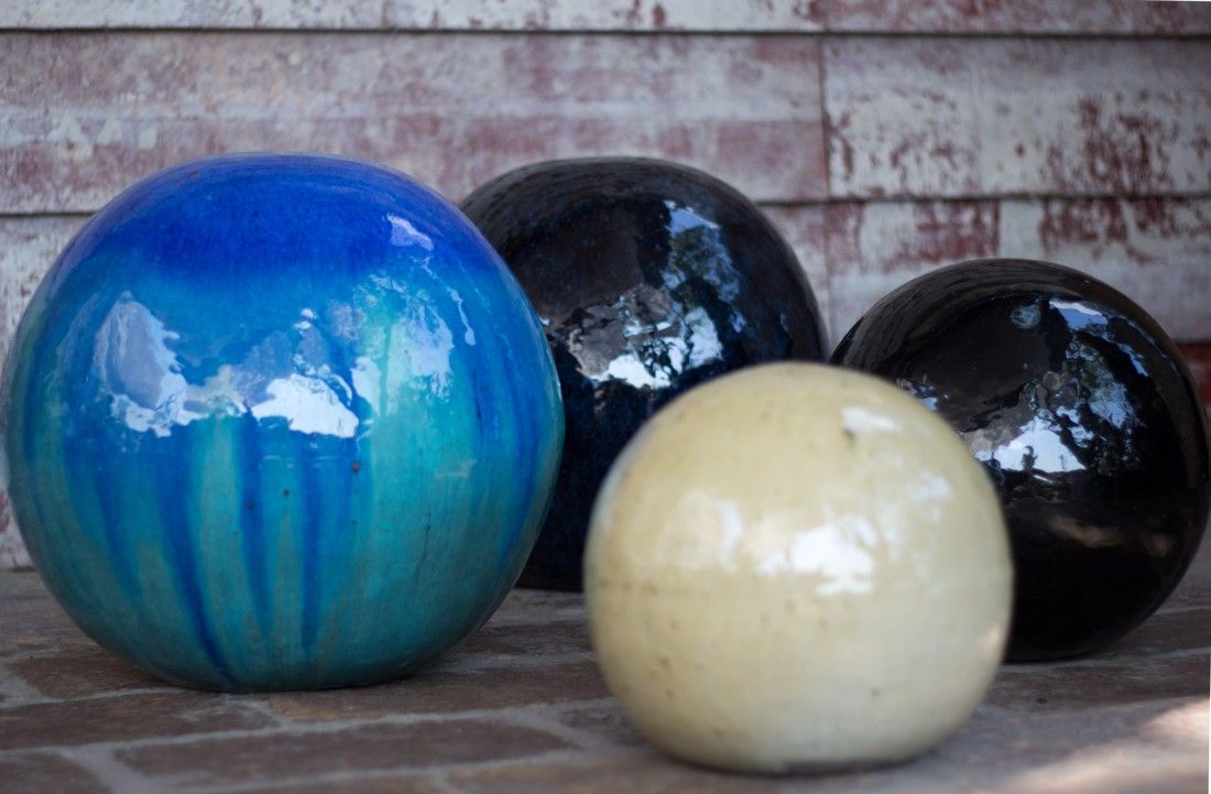 Assorted glazed ceramic garden spheres in blue drip, black, and cream glossy finishes displayed outdoors on a stone patio.