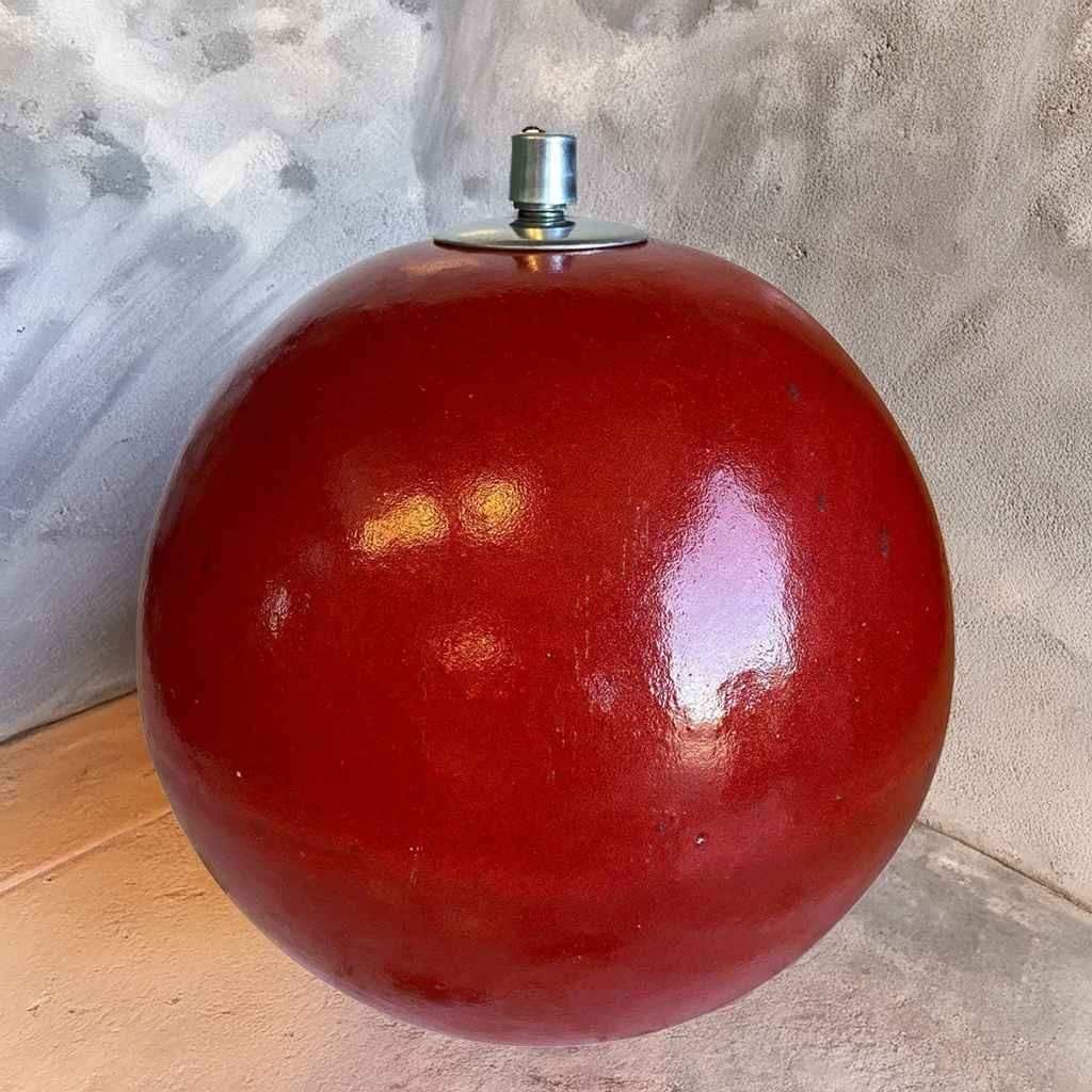 Round red glazed ceramic sphere oil lamp with silver metal wick fixture on top, set against a stone wall.