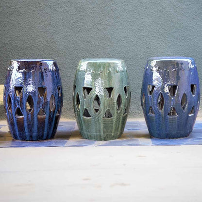 Ceramic garden stools in various glazed hues, including blue and greenish-blue teal, featuring intricate cut-out designs for decorative outdoor seating