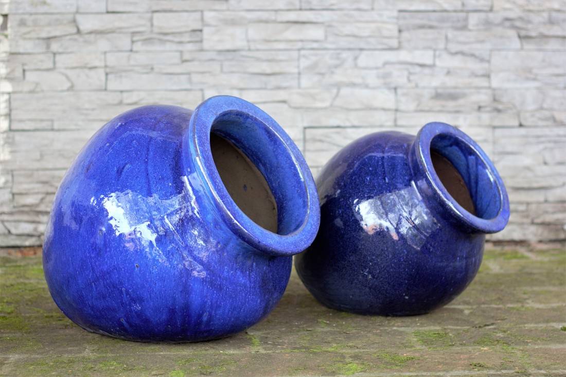 Set of two blue glazed ceramic jar pots with wide round mouths, tilted on a stone surface against a slate wall.