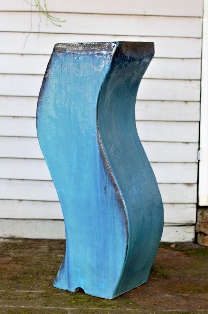 Tall S-shaped curved blue glazed ceramic floor vase against a white wood-paneled wall.