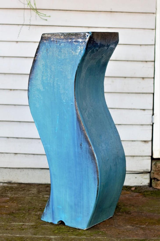 Tall S-shaped curved blue glazed ceramic floor vase against a white wood-paneled wall.