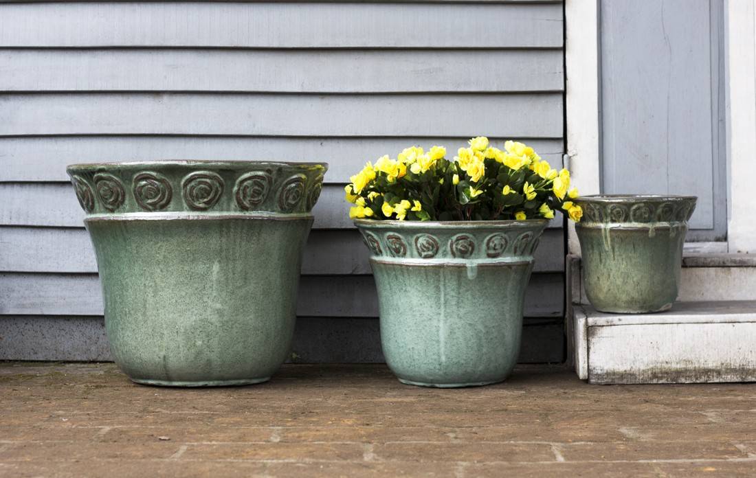 Set of three green glazed ceramic planters with embossed scroll rim detail, one holding yellow flowers, on a porch.