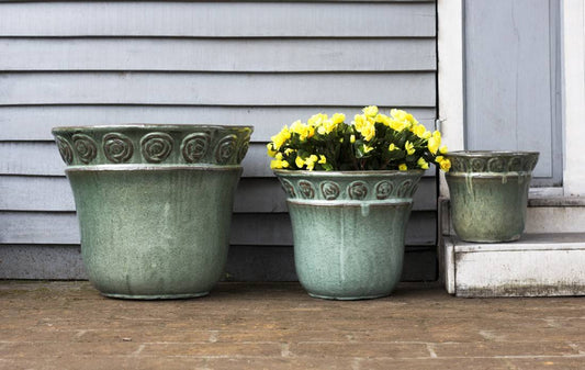 Set of three green glazed ceramic planters with embossed scroll rim detail, one holding yellow flowers, on a porch.