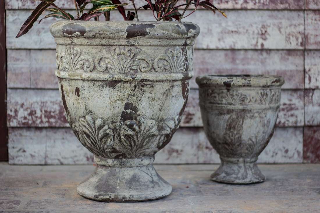 Set of two antique weathered ceramic urn planters with embossed scroll and leaf detail, larger one planted with foliage.
