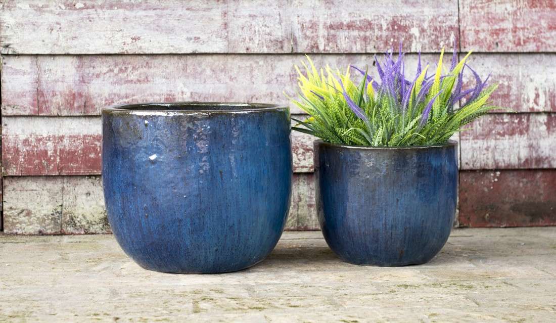 Set of two blue glazed ceramic planters, smaller one holding faux purple and yellow foliage, against a rustic wood wall.
