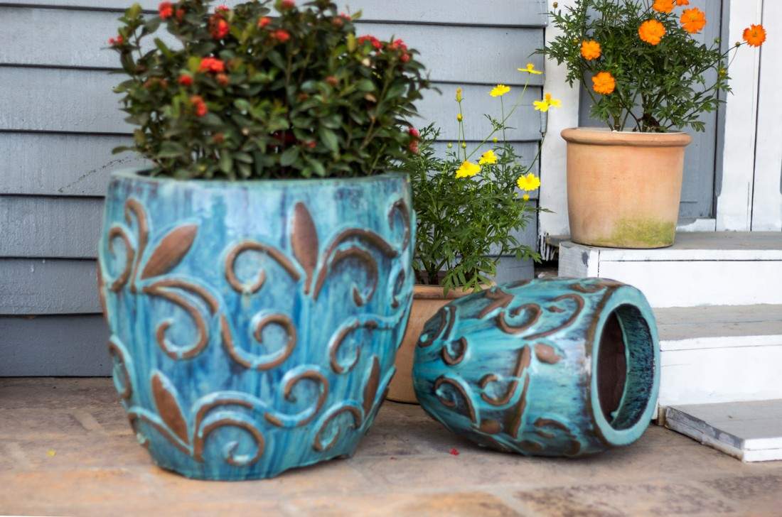 Set of two blue glazed oval ceramic planters with raised scrolled vine relief, one upright with red flowers, one tipped on its side on a patio.