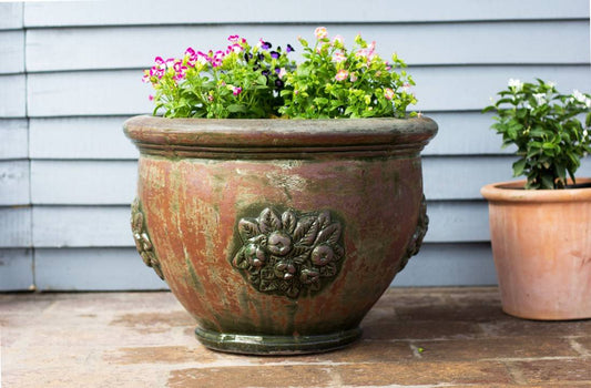 Weathered terracotta ceramic planter with embossed floral and leaf relief, planted with pink and purple flowers, beside a smaller terracotta pot.