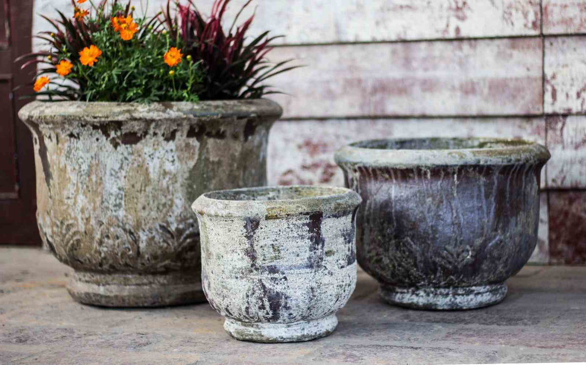 Set of three weathered footed ceramic urn planters with embossed leaf relief, largest one holding orange flowers, on a stone patio.