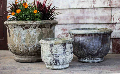 Set of three weathered footed ceramic urn planters with embossed leaf relief, largest one holding orange flowers, on a stone patio.