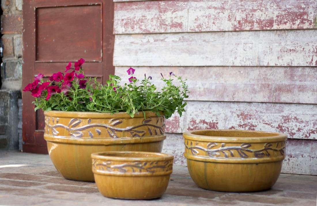 Set of three yellow glazed ceramic planters with embossed vine scroll relief, largest one holding pink and purple flowers, on a porch.