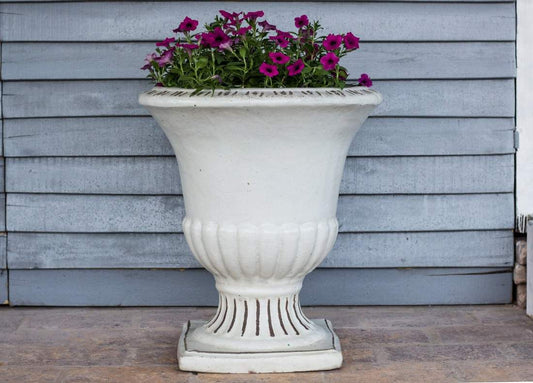 White fluted pedestal urn planter with square base, planted with magenta flowers, against gray wood siding.